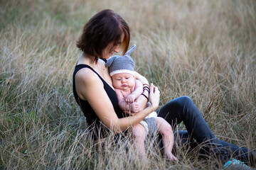 Mother hugging newborn baby boy in her hands outdoors in summer in sun rays, sitting in grass