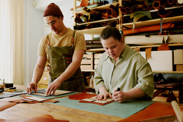 Male tailor and his female colleague with down syndrome cutting out leather details at working...