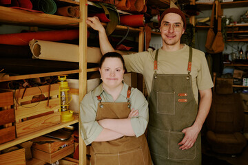 Male and female with down syndrome tailors standing next to wooden shelves full of leather rolls...