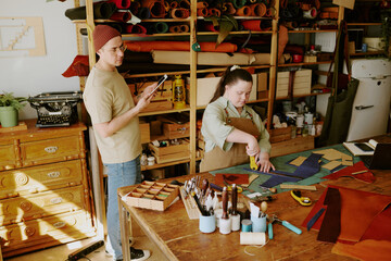 Male tailor holding tablet and controlling his female coworker with down syndrome while she cutting out leather workpieces