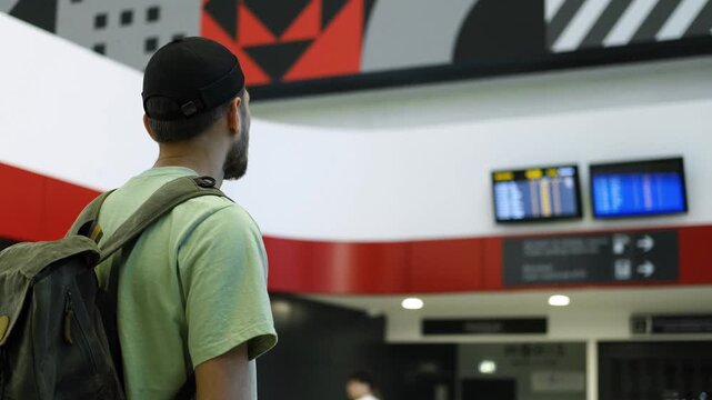 Young man at the airport looking at the list of destinations, rear view. Tourist guy with green retro backpack waits for a train at a train station. A new railway station in Belgrade, Serbia
