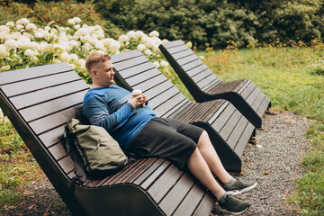 Fototapeta premium Handsome man with backpack sits on a bench and drinks coffee outdoors. Cheerful young man sitting outside on a sunny day, enjoying a relaxed urban setting. Full body