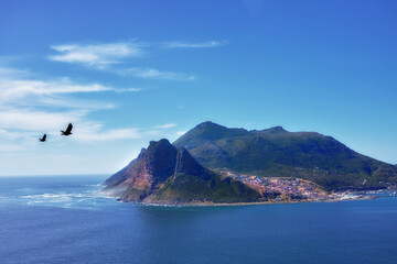 Blue sky, clouds and mountain or ocean with buildings in nature or fishing village by sea. Coastal town, birds and waves or landscape in summer for travel, vacation and outdoor for holiday by beach