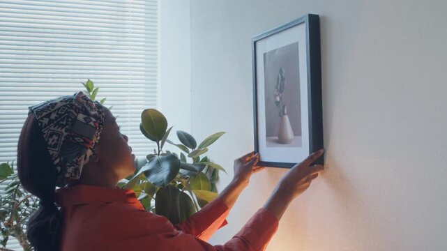 Black young woman hanging frame with picture of vase on white wall standing in corner near window with plants on windowsill