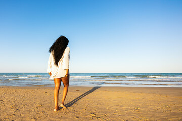 Beautiful African American woman in orange bikini and white shirt on tropical beach. Portrait of dark-skinned woman smiling in sea. Brunette tanned girl in swimsuit enjoying and walking on beach.