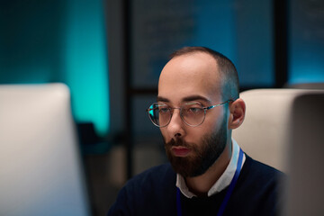 Minimal portrait of bearded computer programmer wearing glasses and looking at screen while working in office with blue light copy space