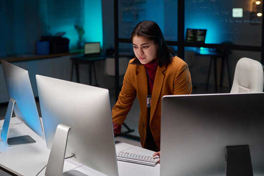 Portrait of female cybersecurity expert using computer with multiple monitors in office lit by blue light