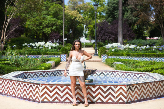 young brunette latin woman with white dress and tattoos in maria luisa park in Seville. The woman is in the fountain of the clam, famous for its romances in the 16th century in Seville, Spain.