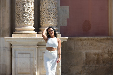 Fototapeta premium Young Spanish woman, brunette and beautiful with white dress in front of the facade of a historic building in the city of Seville, andalusia, Spain. The woman makes different body expressions.