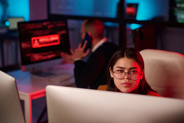 Portrait of young woman wearing glasses and looking at computer screen in cybersecurity department with red emergency light copy space
