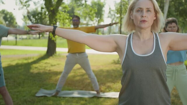 Tilt slowmo shot of middle-aged Caucasian yogini leading outdoor yoga workout, practicing triangle pose with diverse elderly people, training on lawn on warm summer day