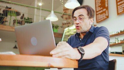 In a cozy cafe, a man with wireless headphones gestures animatedly during a video call on his laptop. Sipping coffee and smiling at the camera, the lush green plants enhance the warm atmosphere.