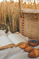 Obraz premium Picnic setup with a woven basket, book, straw hats, croissants, and blueberries on a blanket. Set against a golden wheat field backdrop, capturing a serene countryside atmosphere