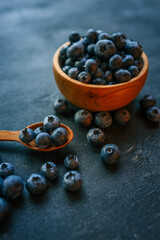 Wooden bowl filled with fresh blueberries on a dark textured background. A wooden spoon lies beside it, with scattered berries creating a rustic and elegant presentation