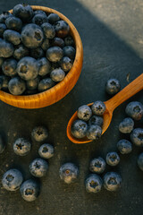 Wooden bowl filled with fresh blueberries on a dark textured background. A wooden spoon lies beside it, with scattered berries creating a rustic and elegant presentation