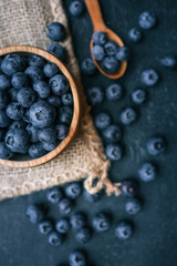 Wooden bowl filled with fresh blueberries and a wooden spoon with a few berries on a dark surface. The rustic presentation highlights the natural beauty and vibrant color of the fruit