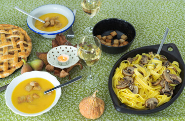 A table with a variety of food, including a pie, soup, and pasta. The table is set with wine glasses and utensils, and there is a candle in a bowl. Scene is warm and inviting