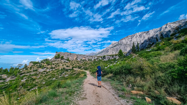 Hiker man with baby carrier backpack on idyllic hiking path to Tucepi town along Makarska Riviera, Dalmatia, Croatia, Europe. Majestic Biokovo mountains rise up against dramatic sky. Dinaric Alps