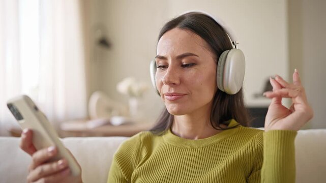 Young Caucasian woman wearing headphones while listening to favourite musical group. Holding smartphone and attentively looking at screen. Messaging with someone and enjoying music.