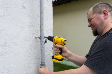A man is using a power drill to secure a pipe to the exterior wall of a home, showcasing his DIY...