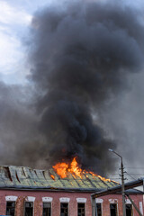 Bright flames erupt from a roof, sending dark smoke billowing into the evening sky as firefighters...