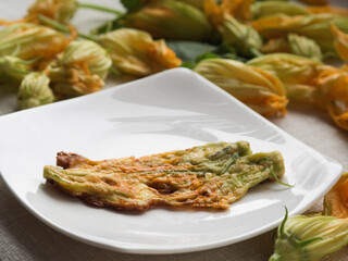 Horizontal Close-Up of Zucchini Flowers Fried in Batter on a White Plate, Surrounded by Fresh Zucchini Flowers on a White Tablecloth.