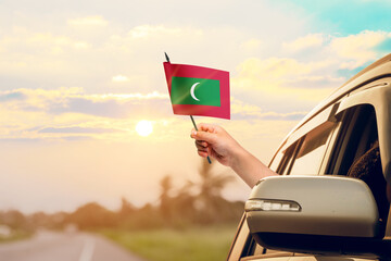 Waving the Maldives flag against the sunrise or sunset from a car driving along a country road. Holding the Maldives flag, traveling by car, on a weekend trip.