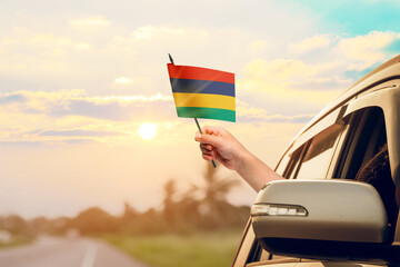 Waving the Mauritius flag against the sunrise or sunset from a car driving along a country road. Holding the Mauritius flag, traveling by car, on a weekend trip.