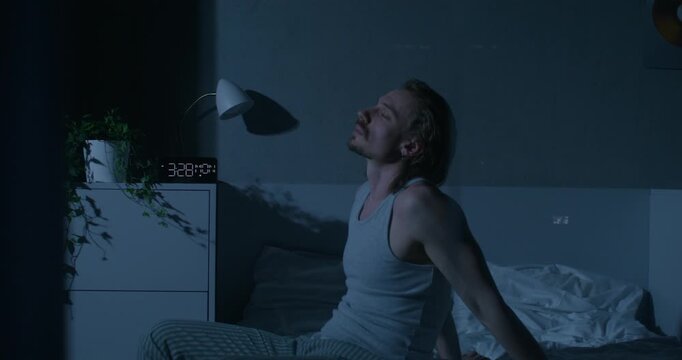 A man rubs his face while sitting on the bed, he can't sleep. Home interior, dark room