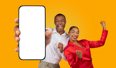 African American happy couple standing against a yellow background. The man is holding a smartphone with a blank screen in front of him and pointing at it with his right hand.