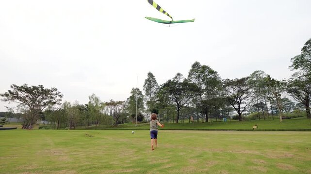 Little boy runs and accelerating as his kite with striped tail flies behind him. Shot in slow motion, toddler runs away, seen from rear view. He spots soccer ball, kicks it, and continues running