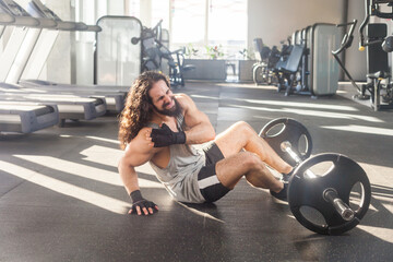 Portrait of grimace muscular man with long curly hair working out in gym, sitting on floor injured arm, spasm painful, touching his shoulders frowning face. Indoor shot.