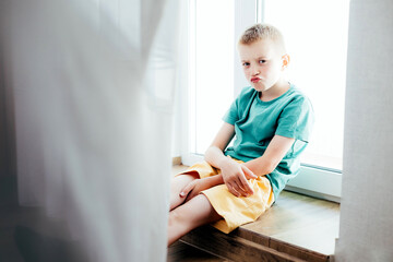 Cheerful school-age boy in a green T-shirt sitting at the window at home. Front view