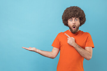 Wow, look at commercial proposal! Portrait of man pointing at copy space on palm and looking with amazement, showing empty place for idea presentation. Indoor studio shot isolated on blue background.