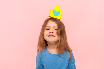 Portrait of funny little girl standing with small paper house on her head, looking at camera, relocating, renting, wearing blue sweater. Indoor studio shot isolated on pink background.