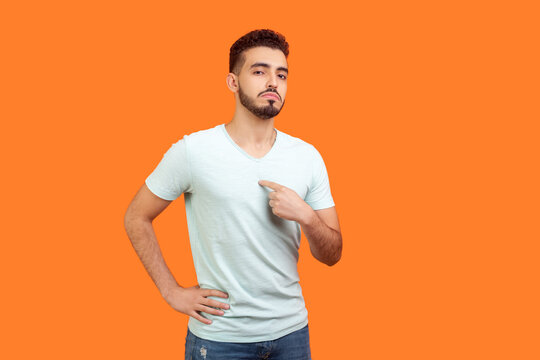 Portrait of proud confident attractive young bearded man wearing T-shirt standing pointing at himself looking at camera with bossy face. Indoor studio shot isolated on orange background.