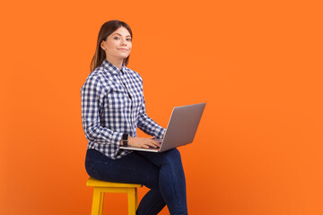 Naklejka premium Portrait of proud executive woman with brown hair working on laptop online, distance job on computer, wearing checkered shirt. Indoor studio shot isolated on orange background
