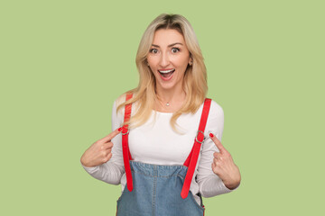 Portrait of amazed excited adult blond woman standing pointing at herself with amazement, looking at camera, wearing denim overalls. Indoor studio shot isolated on light green background