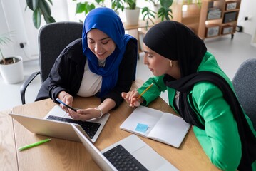 Two women in hijabs engaging in a collaborative brainstorming session at a modern office, using laptops and writing notes.