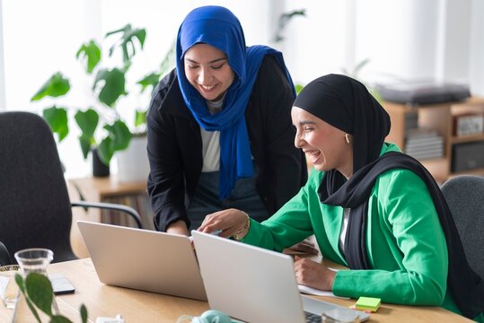 Two women in hijabs collaborating on a project in a modern office, using laptops and sticky notes to outline their strategies.