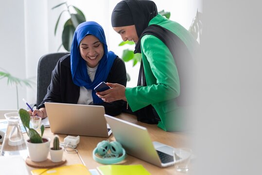 Two women in hijabs focusing on goal-oriented team planning in a modern office, using laptops and sticky notes to set their objectives