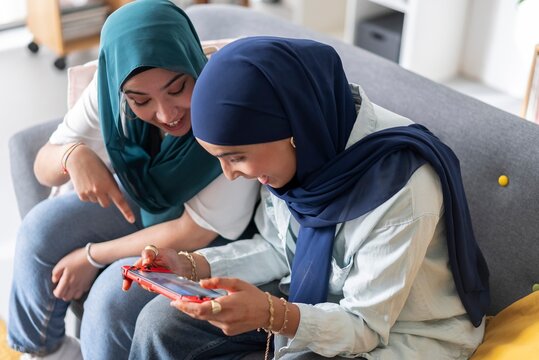 Two women in hijabs enjoying a gaming session on a handheld console, sharing smiles and laughter, creating a joyful and engaging atmosphere in a modern living room