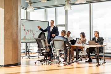 A group of people is sitting around a table in a modern office, listening to a presentation on a...