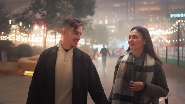 Happy young couple in trendy clothes talking while walking on footpath on Christmas night. Loving partners strolling in illuminated city decorated with string lights. Winter holiday season.