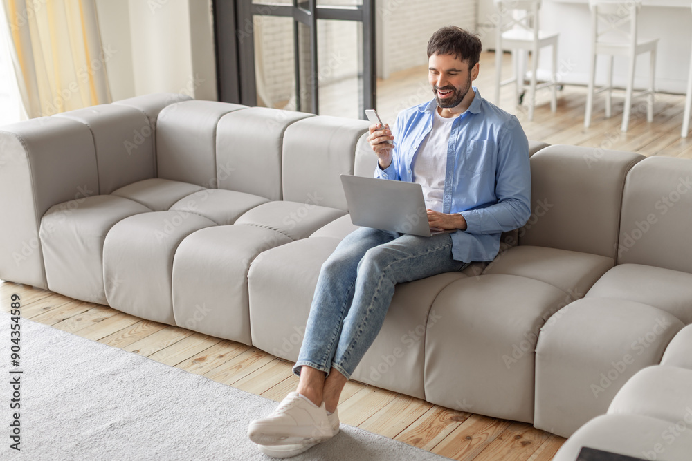 Poster a man sits on a modern gray sectional sofa in a living room. he is looking at a smartphone and smili