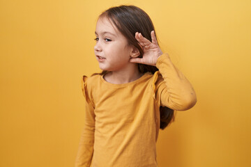 Adorable hispanic girl tuning in! standing over yellow, isolated background, hand cupped over ear,...