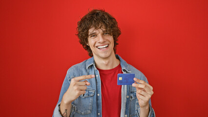 Happy young man with curly hair pointing at credit card against vibrant red background