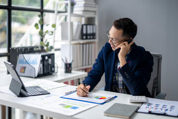 Fototapeta premium A man is talking on his cell phone while sitting at a desk with a laptop
