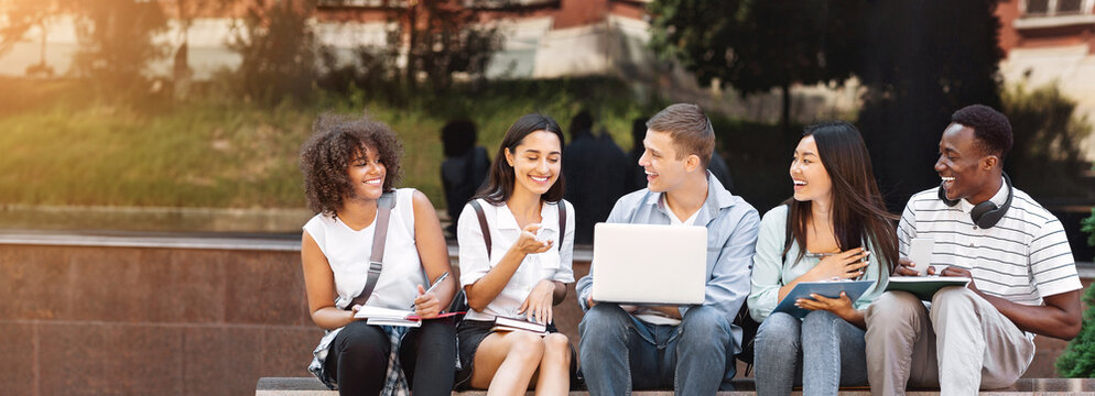 Campus Life. Cheerful college students resting outdoors between classes, using laptop, chatting and laughing, free space