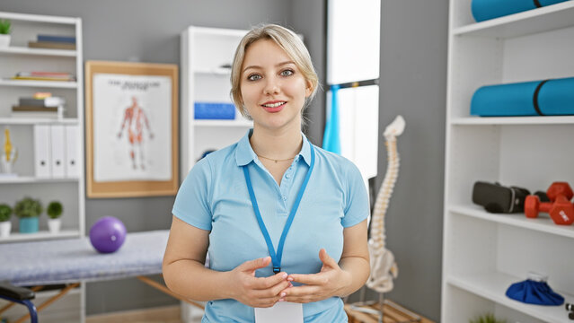 A professional young woman stands confidently in a rehabilitation clinic, surrounded by anatomical charts and physiotherapy equipment.
